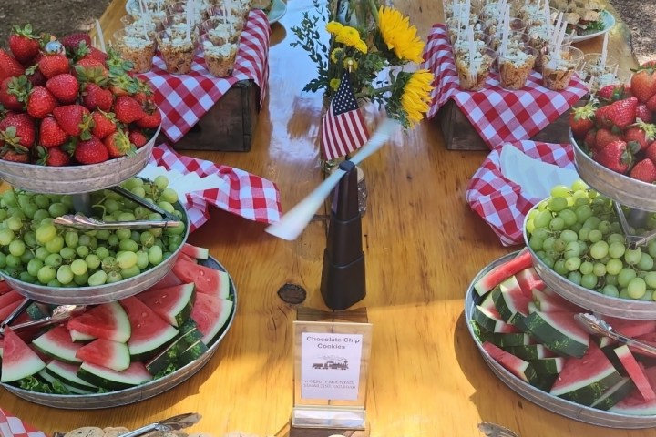 Festive outdoor table with fruit platters, cookies, sunflowers, and checkered napkins.