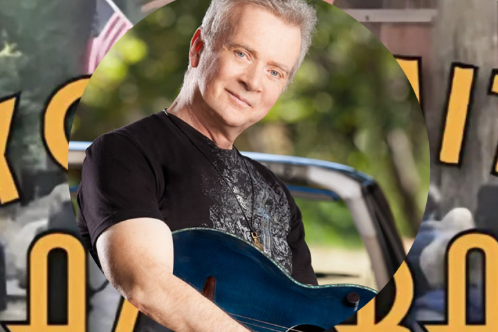 Man holding a guitar in front of a blurred outdoor scene.
