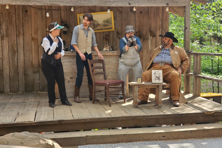Four actors in western attire perform a scene on a wooden stage.