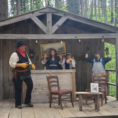 Three performers on a rustic wooden stage, one framed like a painting, others acting out a scene.
