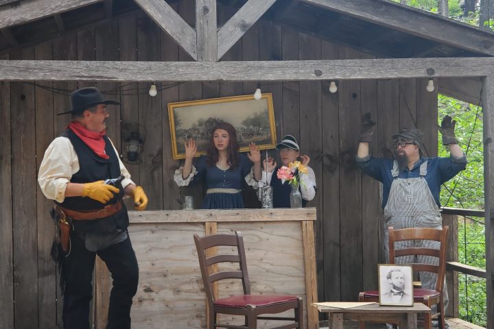 Three performers on a rustic wooden stage, one framed like a painting, others acting out a scene.