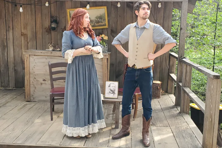 Two people in period clothing on a wooden porch, with a picture on a chair between them.