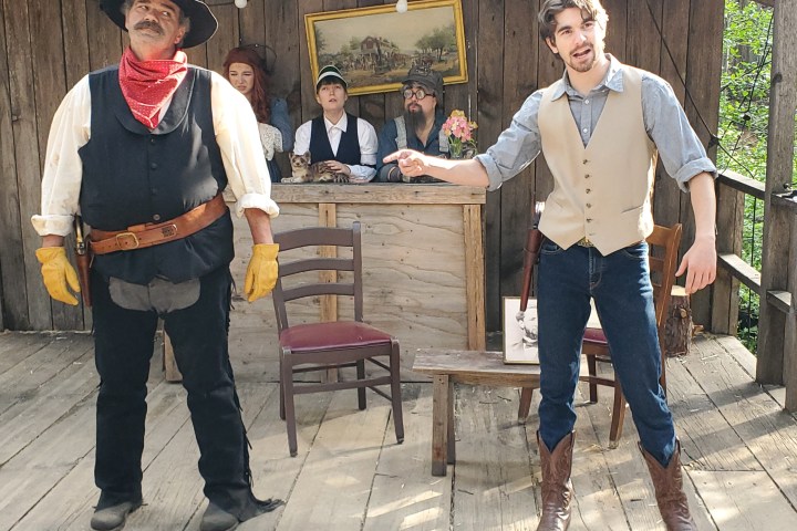 People in cowboy attire perform in a wooden saloon set with props and costumes.